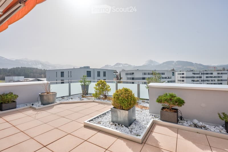 Modern rooftop terrace with tiled flooring, white gravel planters, and potted shrubs, overlooking a cityscape with modern buildings and distant mountains.