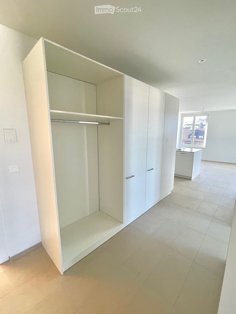 Empty room with white walls, tiled floor, large wardrobe with mirror, rack inside, white kitchen area visible, windows with view of mountains.