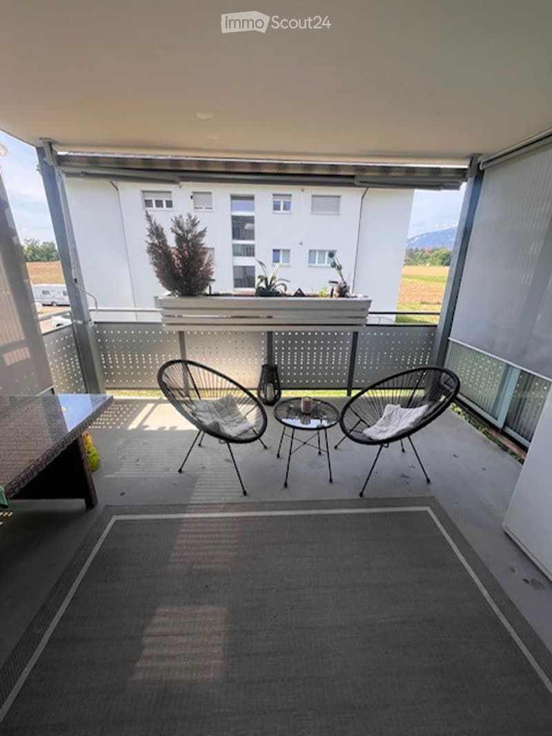 Balcony with two modern chairs, a small table, and a potted plant, overlooking a scenic view of a building and countryside.
