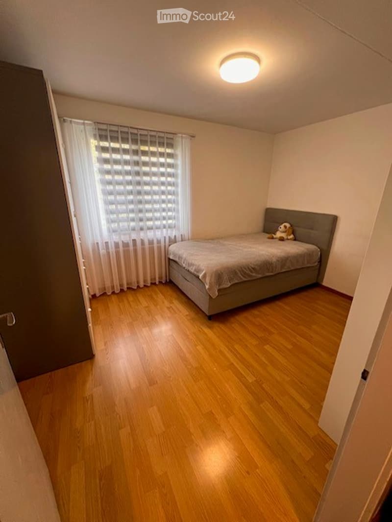 Cozy bedroom with wooden flooring, a grey bed with a grey duvet, a small stuffed toy on the bed, and a window with white curtains.