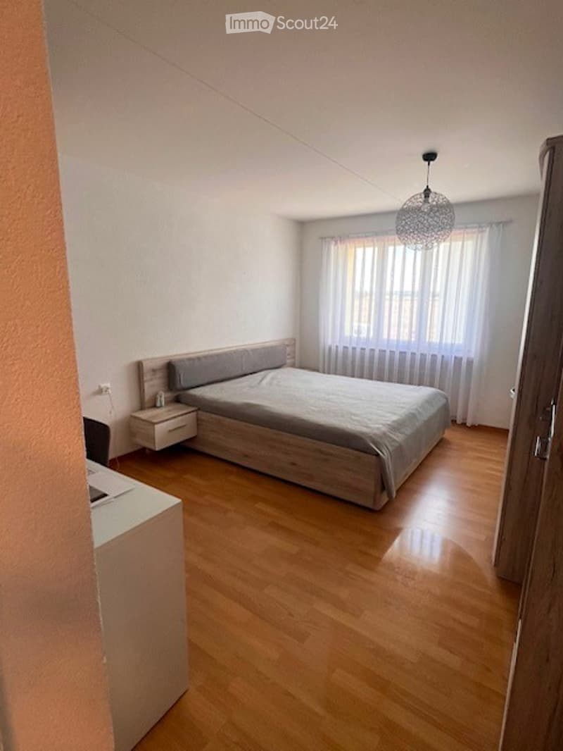 Modern bedroom with wooden flooring, a large bed with grey bedding, a nightstand, and a hanging light fixture.