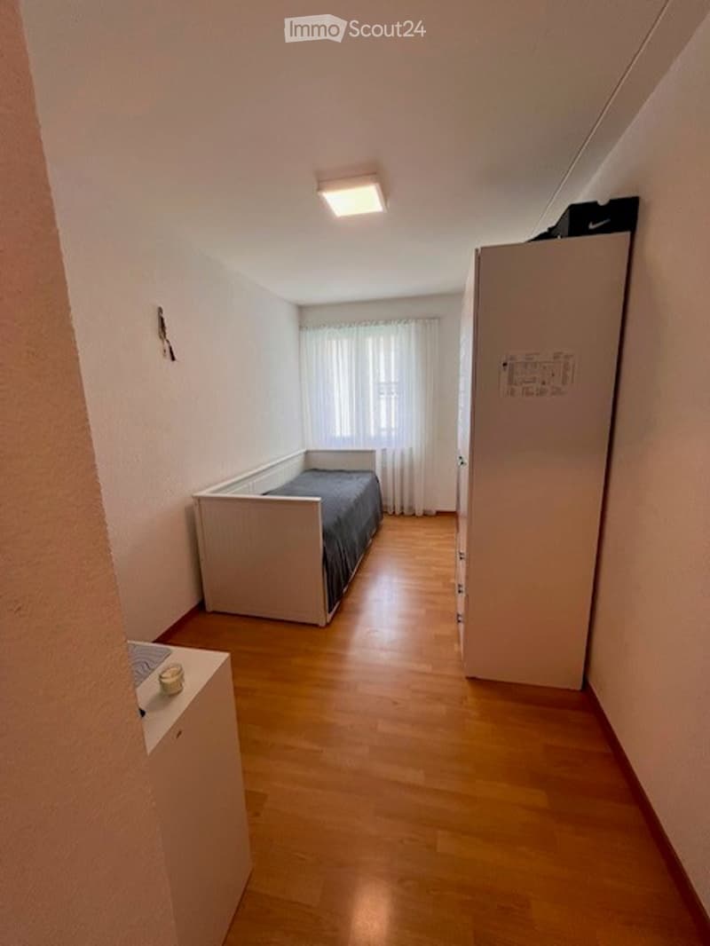 Small furnished room with wooden flooring, a single bed, a nightstand, a radiator, and a cabinet.