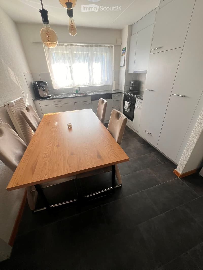 Modern kitchen with white cabinets, dark countertops, a wooden dining table with chairs, and various appliances including a coffee maker and oven.