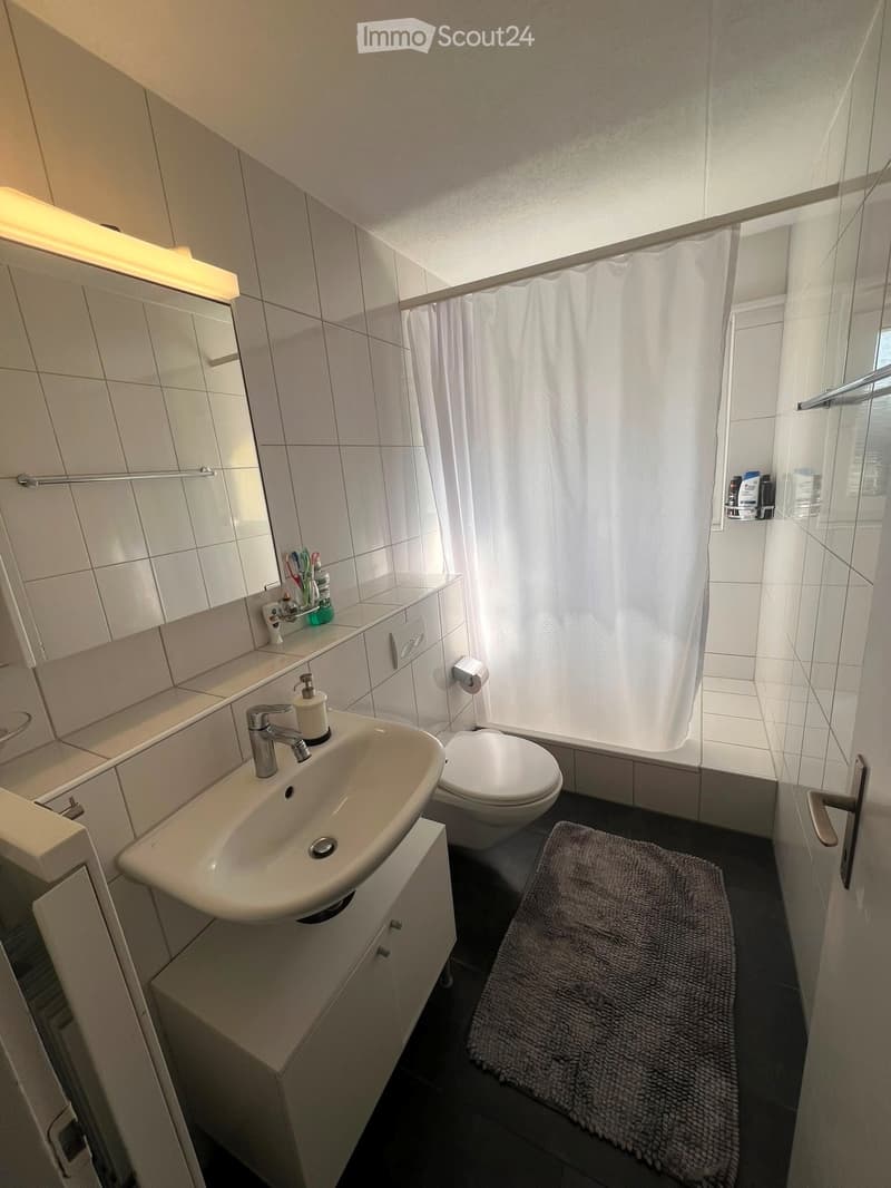 Modern bathroom with white tiles, a sink with a modern faucet, a toilet, a shower with a curtain, a mirror, a bathroom cabinet, and a rug on the floor.