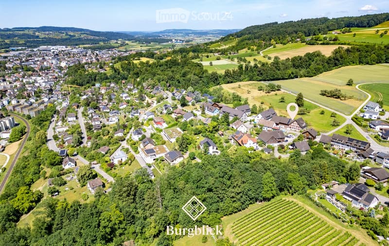 Aerial view of a residential area with numerous houses, surrounded by green fields, trees, and distant hills.