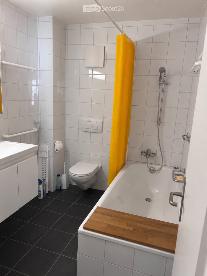 Modern bathroom with white tiled walls, black tiled floor, bathtub with wooden tray, toilet, sink, and shower curtain.