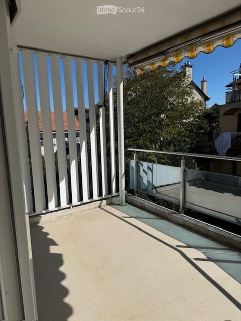 Balcony with white slatted privacy screen, glass railing, and striped awning. The view shows neighboring houses and greenery.