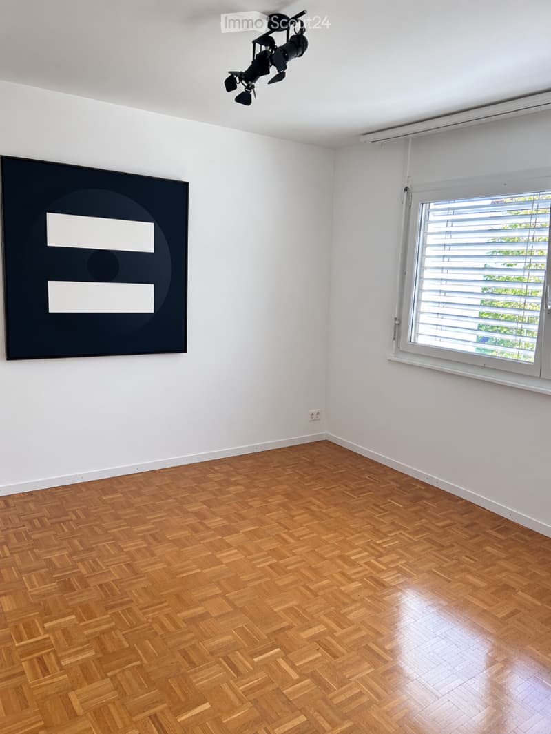 Empty room with wooden parquet flooring, white walls, large window with blinds, and a modern black artwork on one wall.