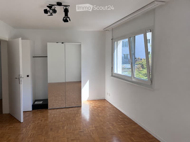 Empty room with wooden flooring, large window, mirrored closet, and track lighting.