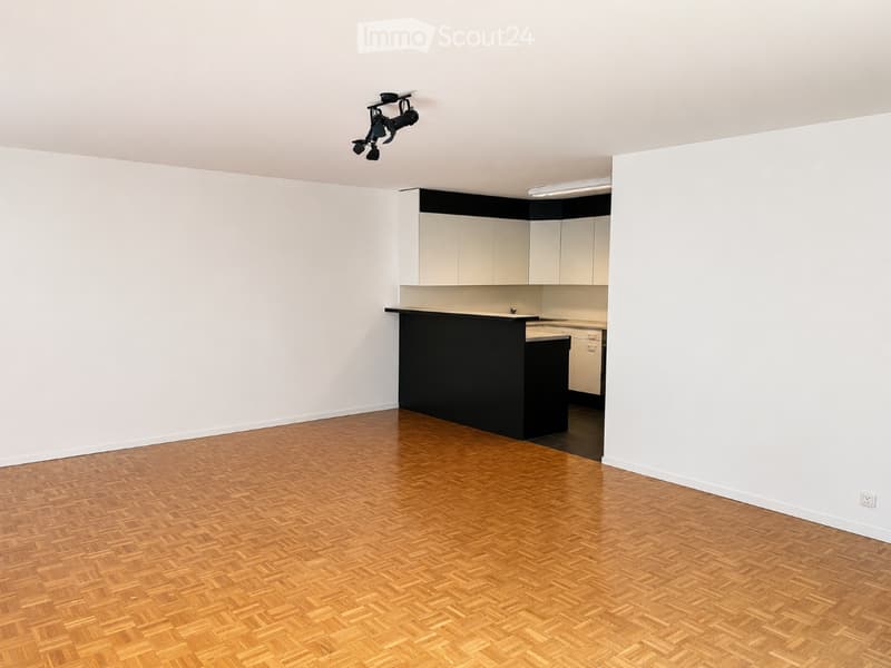 Empty room with wooden herringbone flooring, white walls, and a small kitchen area with white cabinets and a black countertop.