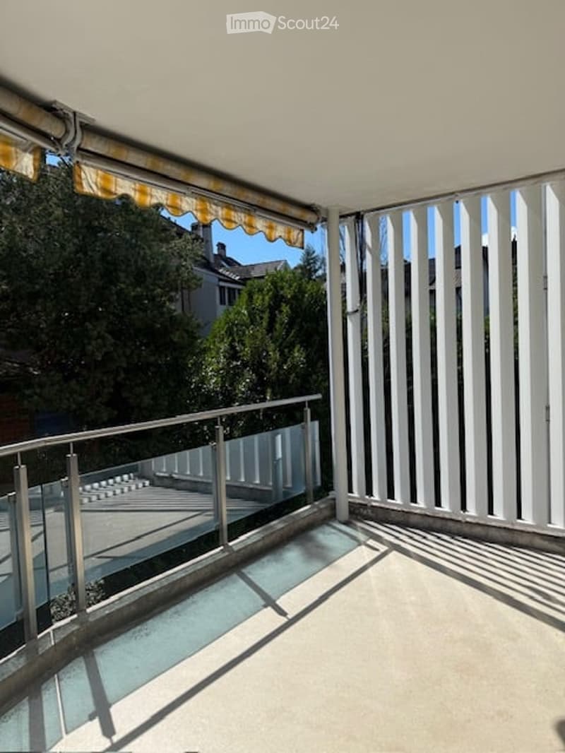 Balcony with white slatted privacy screens, metal railing, and retractable awning. Offers a view of greenery and neighboring buildings.