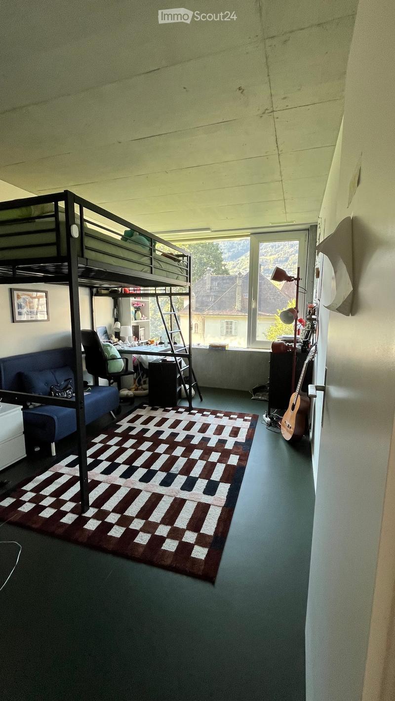 A furnished room with a bunk bed, desk, sofa, chair, and guitar, featuring a patterned rug and large windows with a scenic view.