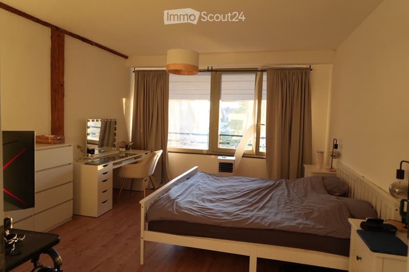 Cozy bedroom with a double bed, wooden floor, large window with curtains, desk with chair, mirror, and nightstand.