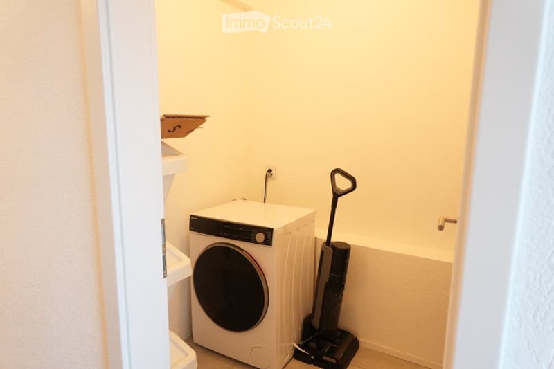 Small bathroom with a white washing machine, a vacuum cleaner, and a shelf above the sink.