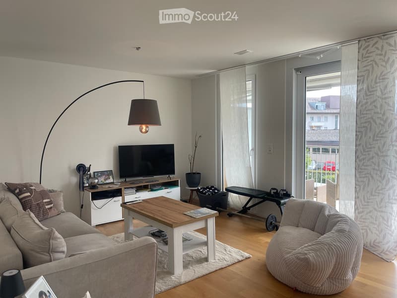 Modern living room with a grey sofa, wooden coffee table, armchair, TV, floor lamp, and large windows with sheer curtains.