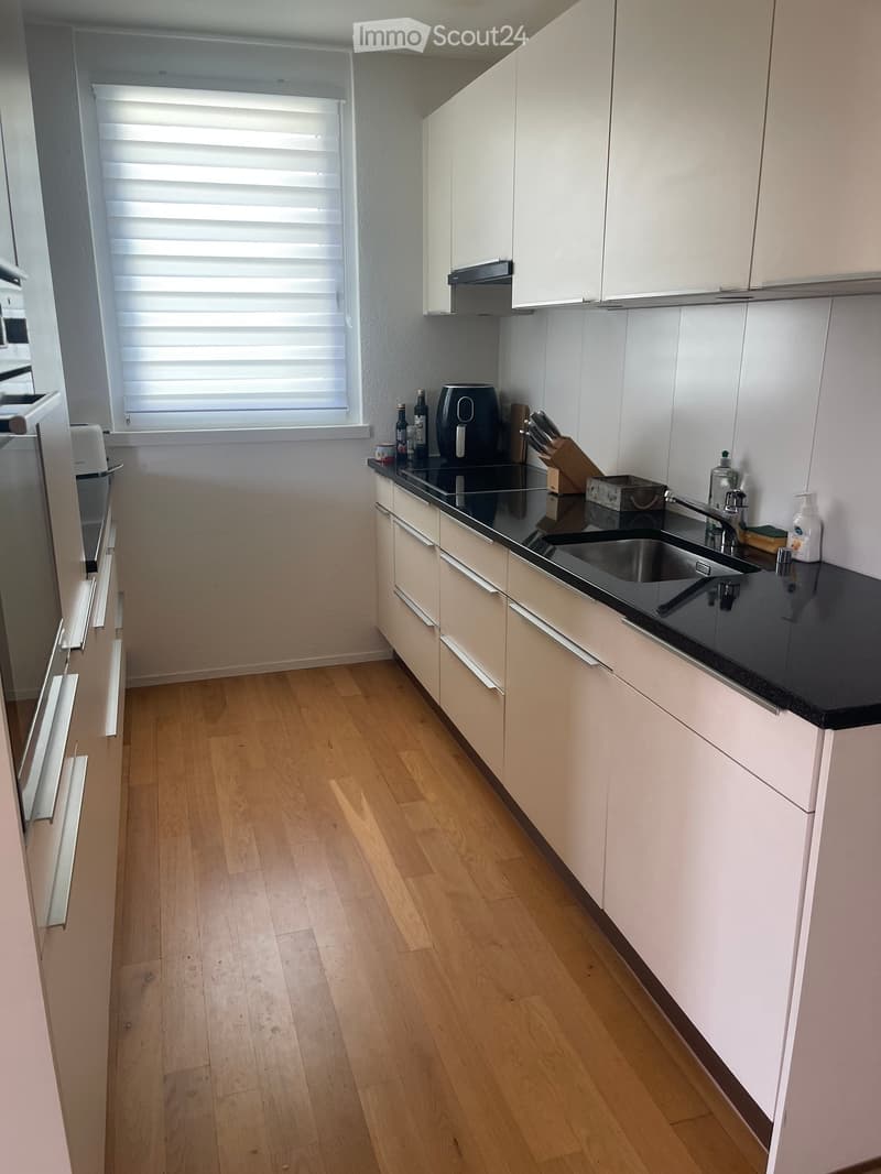 Modern kitchen with light-colored cabinets, black countertop, wooden floor, and various appliances including a fridge, sink, and air fryer.