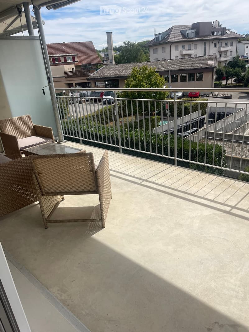 Balcony with wicker chairs and a glass table, overlooking a residential area with buildings and parked cars.