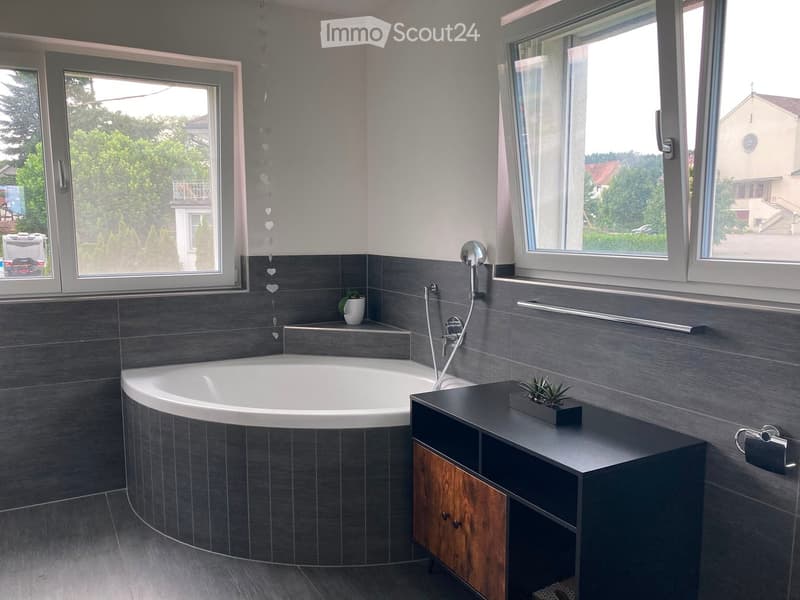 Modern bathroom with a large, curved bathtub, a dark grey tiled wall, a wooden vanity with a sink, and a small potted plant on the counter. The room features large windows with a view of greenery outside.