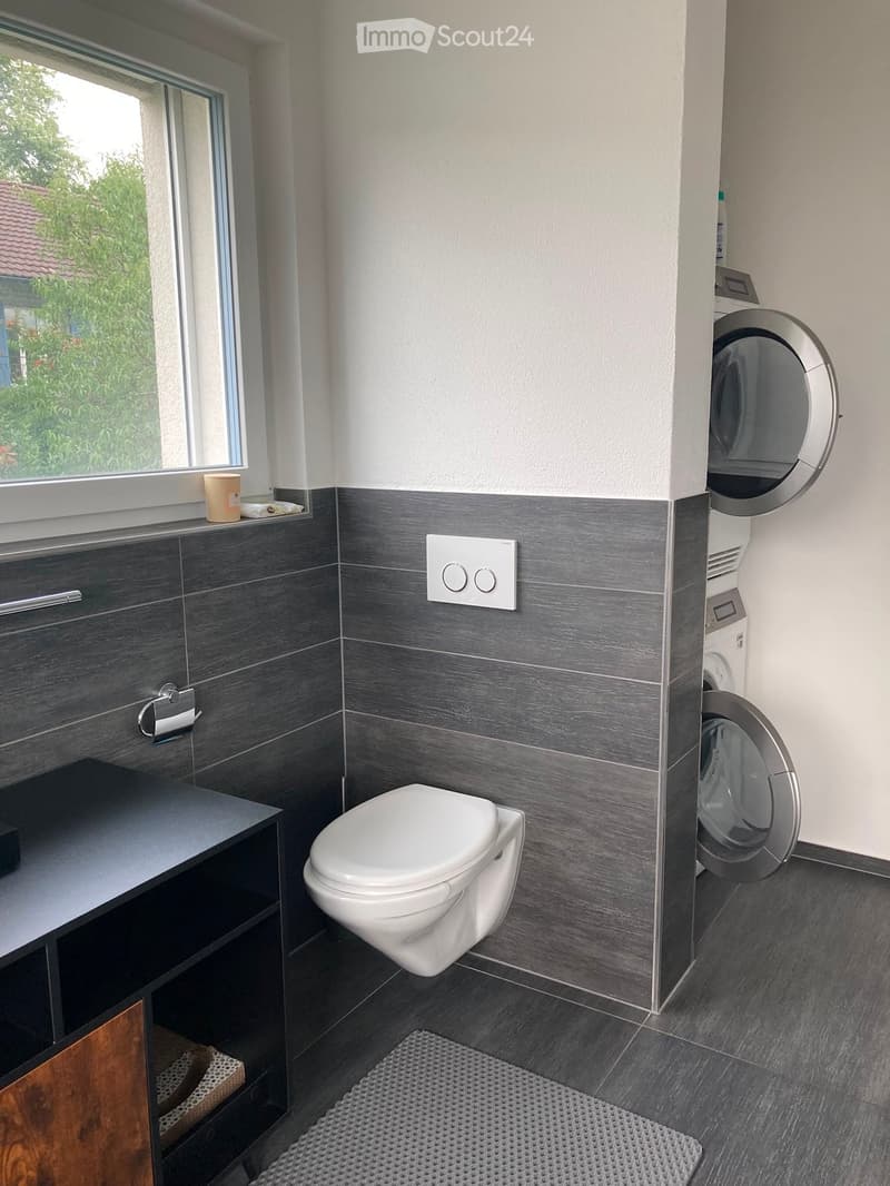 Modern bathroom with dark grey tiles, white toilet, washing machine, and a small shelf with a cup on the windowsill.