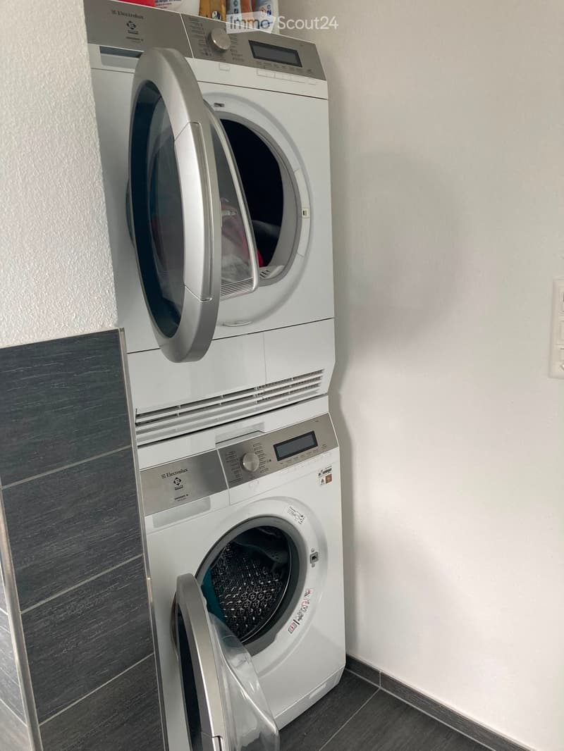 A compact bathroom with a stacked washing machine and dryer unit, both white with control panels, placed against a white wall with grey tiled flooring.