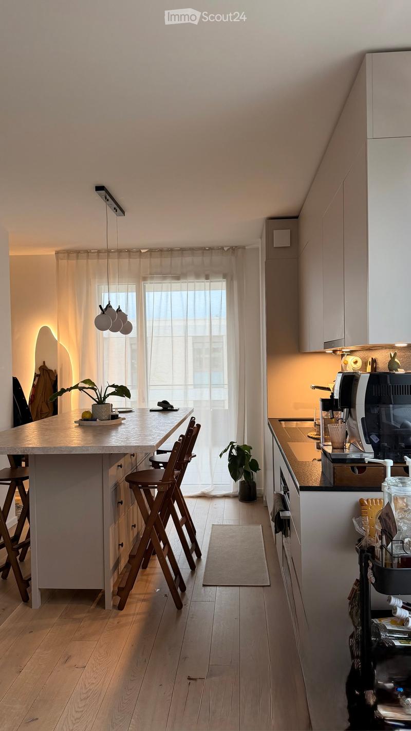 Modern kitchen with a central island, bar stools, coffee machine, and various appliances. The space features wooden flooring, white cabinets, and a large window with sheer curtains.