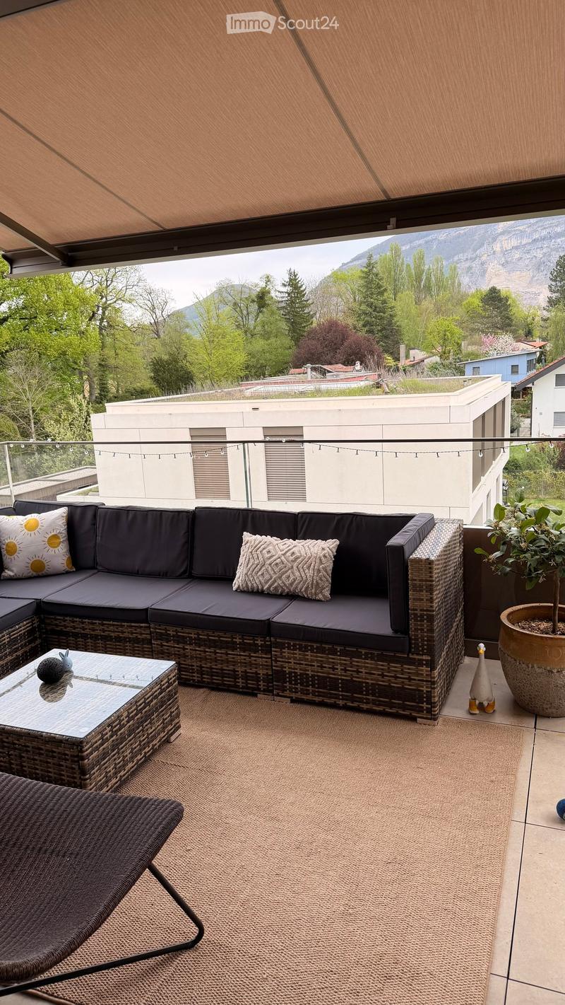 Balcony with a wicker sofa, coffee table, and potted plant, overlooking a scenic mountain view.