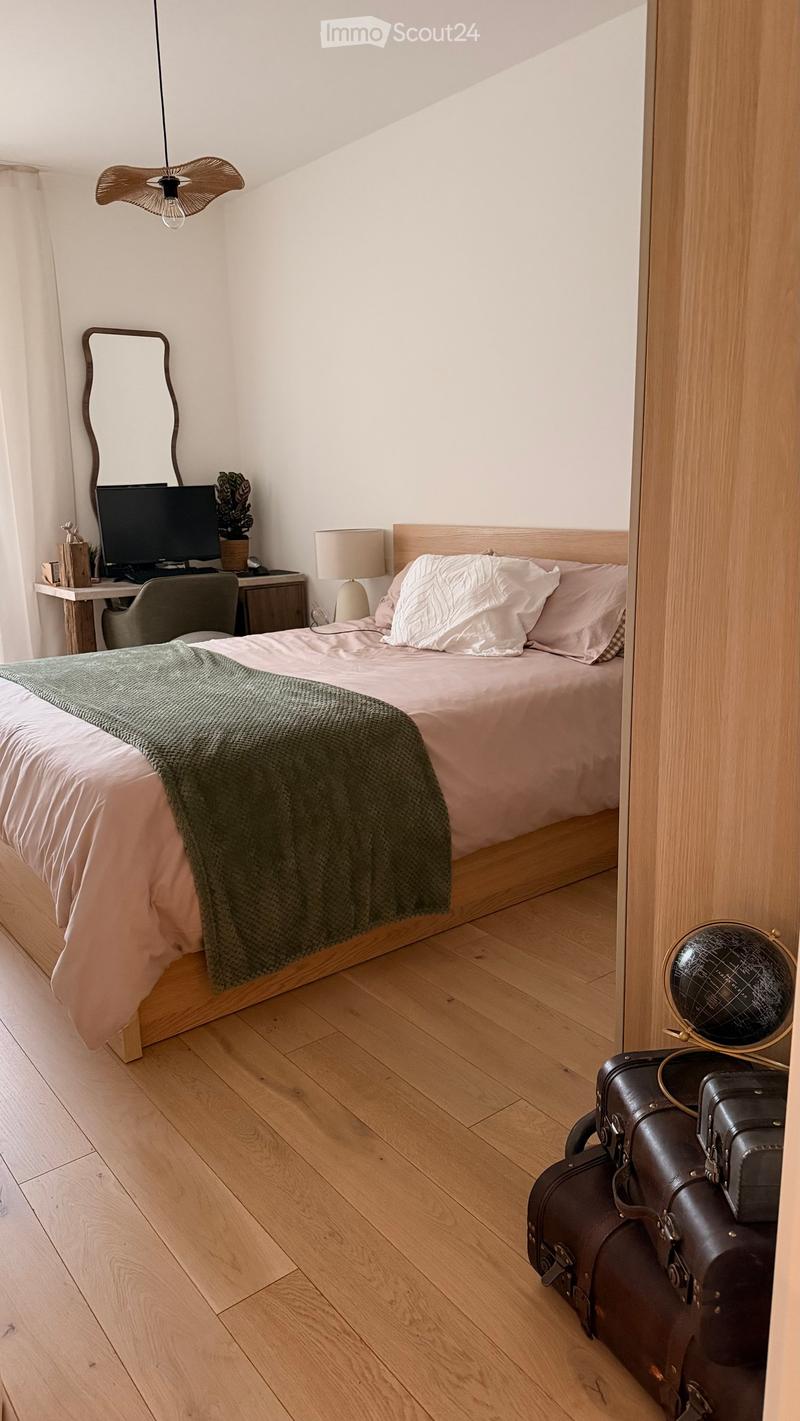 Cozy bedroom with wooden flooring, a neatly made bed with pillows and a green throw, a desk with a chair, a mirror, and a globe on a stand. Suitcases are placed near the door.