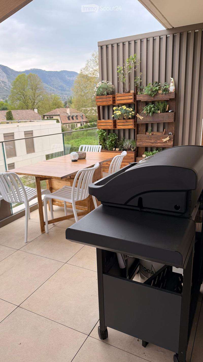 Balcony with a wooden table, white chairs, a grill, and several potted plants, overlooking a scenic mountain view.