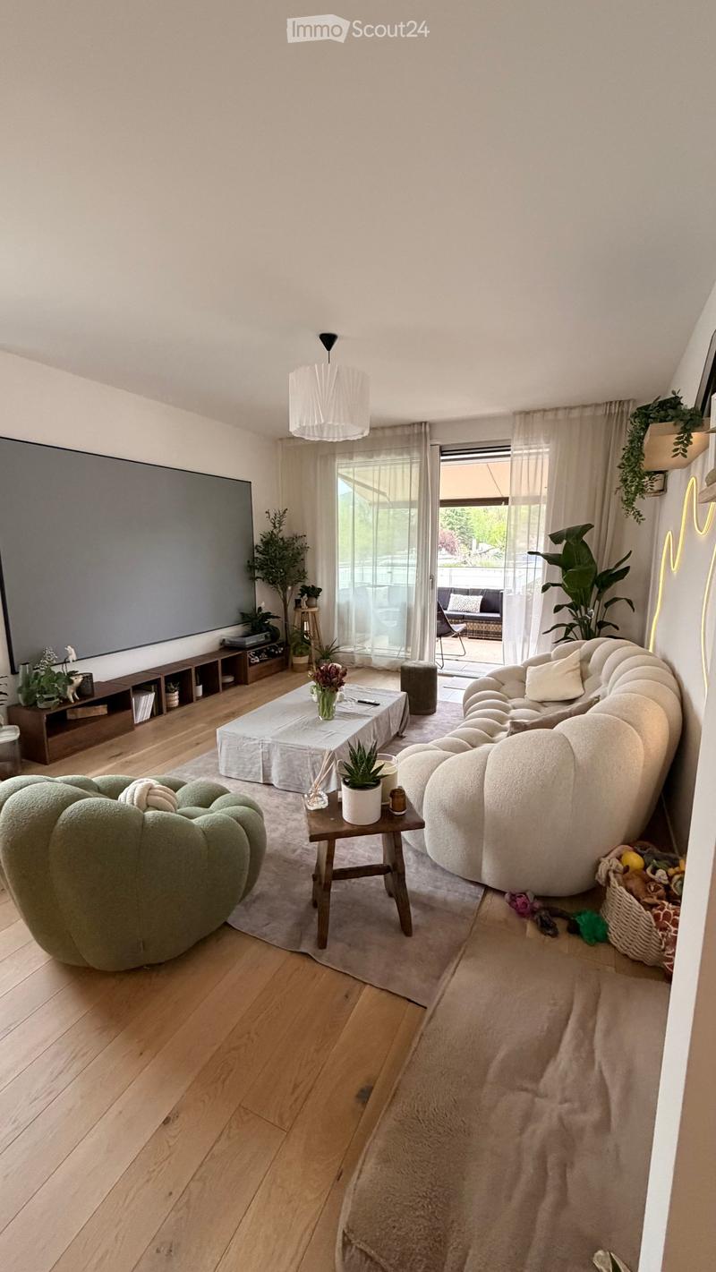 Modern living room with wooden flooring, large window, white sofa, coffee table, ottoman, side table, potted plants, and a TV screen.