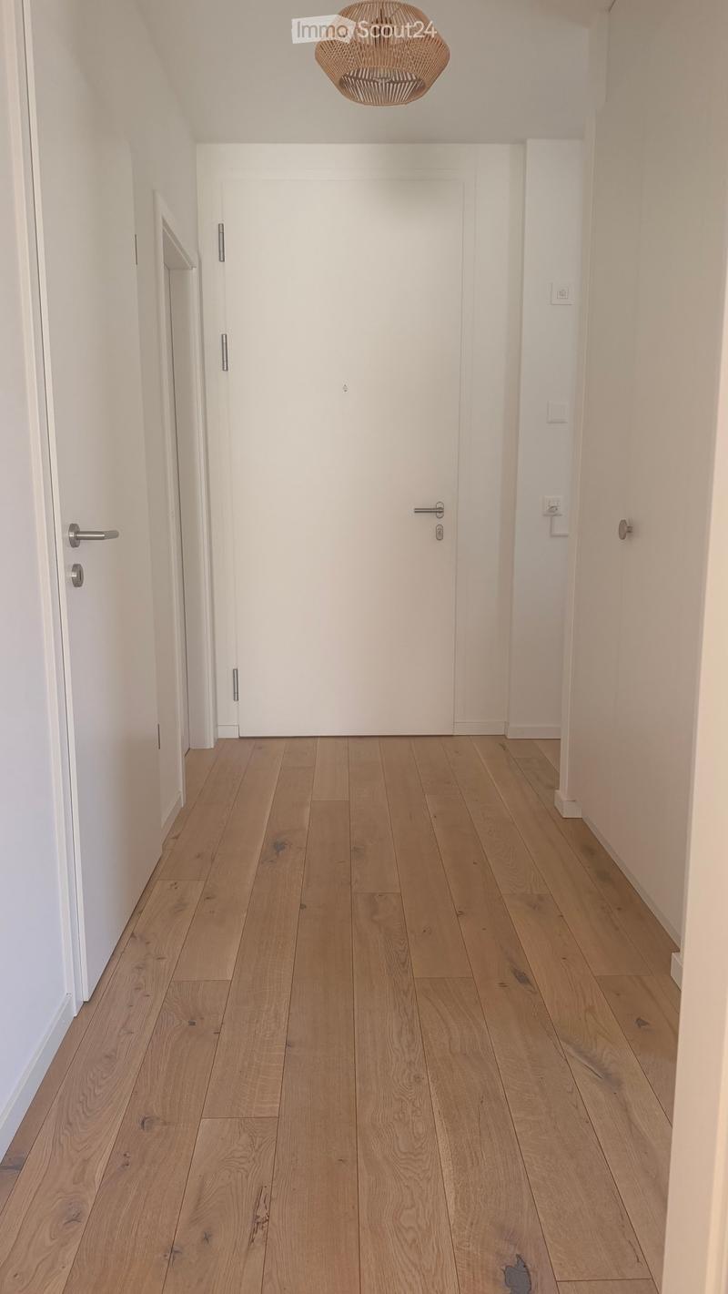 A clean, minimalist hallway with light wooden flooring, white walls, and a single hanging woven light fixture. Two white doors are visible, one on each side.