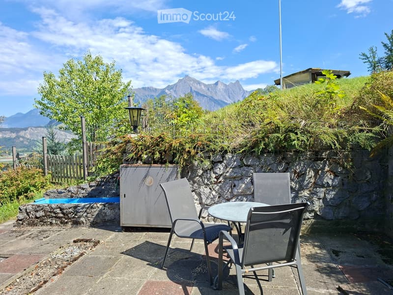 Outdoor patio with a table and chairs, surrounded by greenery and a scenic mountain view.
