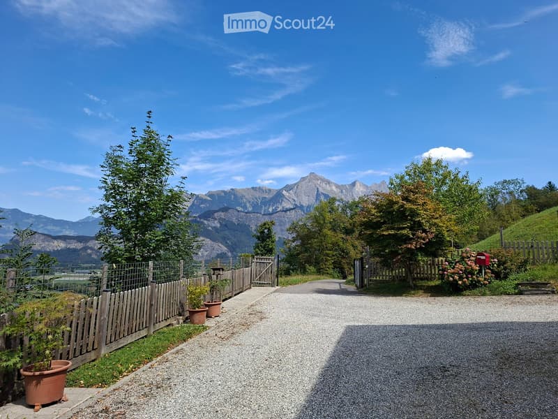 A gravel driveway leads up to a wooden gate, flanked by wooden fences and potted plants, with a scenic mountain backdrop.