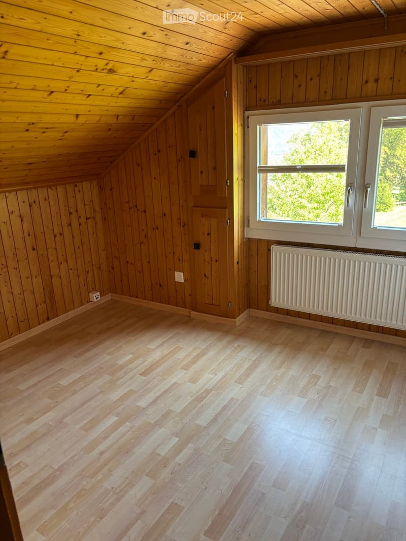 Empty wooden room with slanted ceiling, wooden walls, hardwood floor, and large windows with white frames.