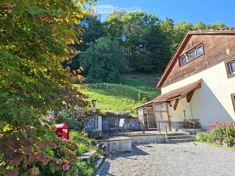 A single-story house with a wooden exterior and a sloped roof, situated on a raised ground floor with a stone retaining wall and steps leading up to a patio area with outdoor furniture.