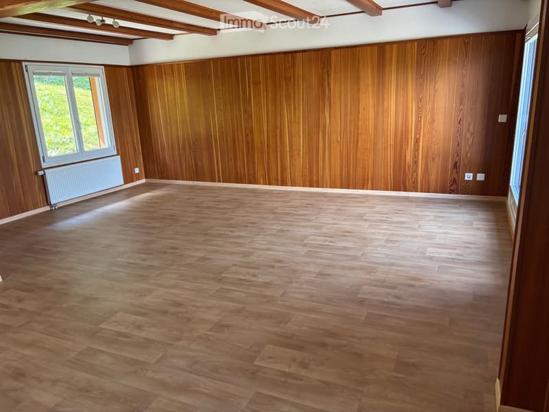 Empty room with wooden walls and floor, large windows, exposed wooden ceiling beams, and a radiator on one wall.