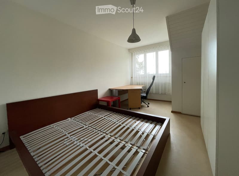 A small, unfurnished room with a built-in bed frame, a desk, a chair, and a small red stool. The room has light-colored walls, a window with white curtains, and a hanging light fixture.