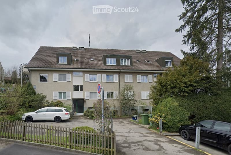 Multi-story residential building with beige facade, multiple windows, and a small garden area. Two cars are parked in front.
