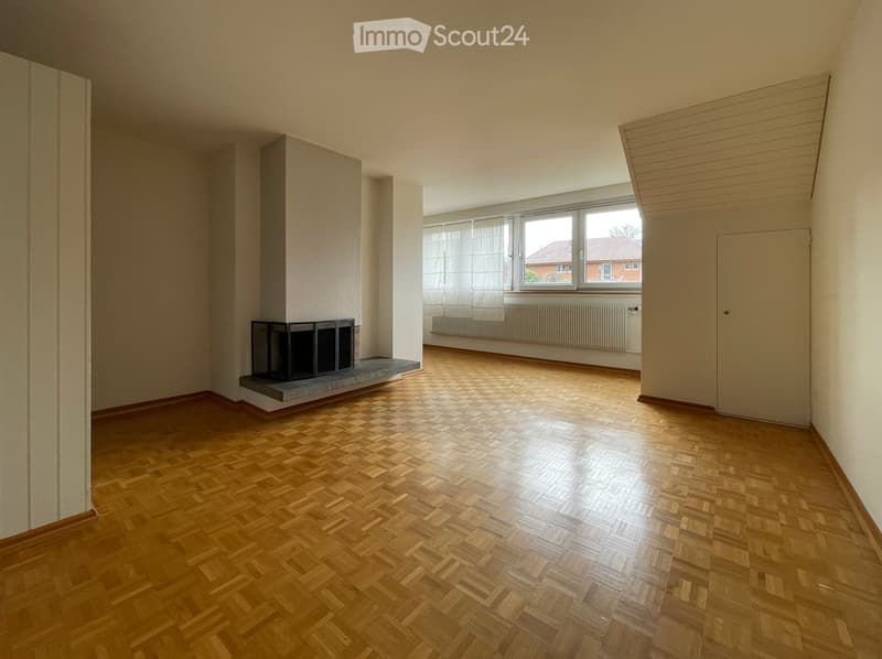 Empty room with wooden herringbone flooring, white walls, and a fireplace with a black metal surround. Large window with white blinds and radiator below.