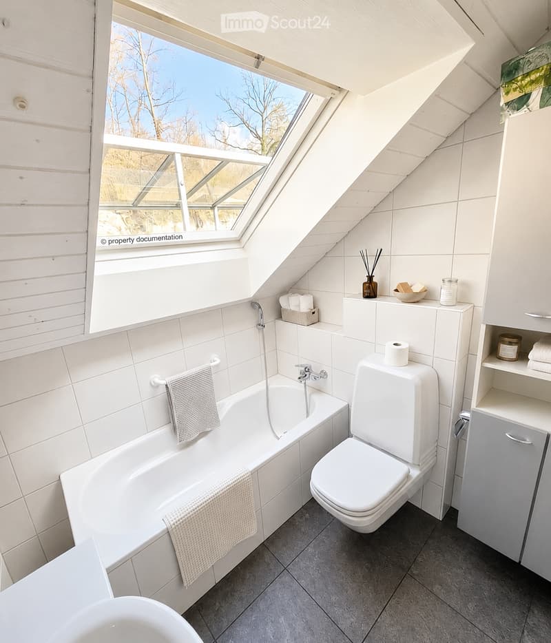 Modern bathroom with a bathtub, toilet, and a large skylight window. White tiles, grey towels, and a small shelf with decorative items.