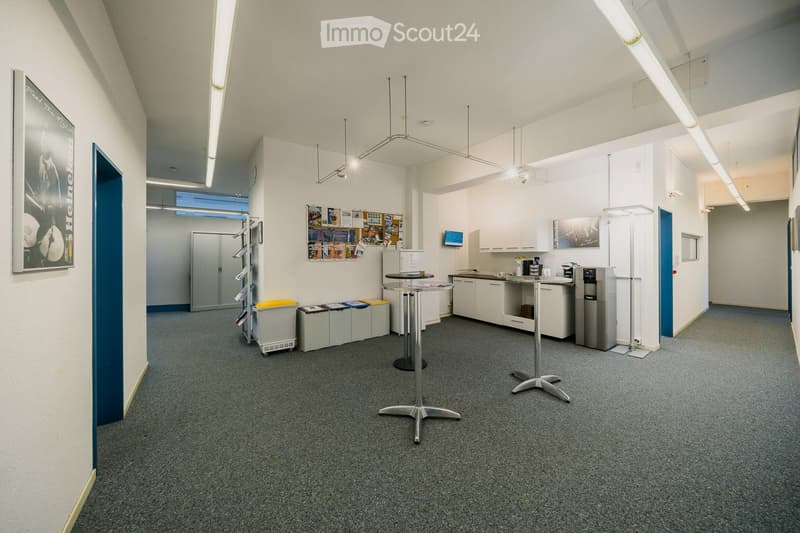 A modern break room with grey carpeting, white walls, and a mix of furniture including tables, cabinets, a screen, and a lamp. There are posters on the walls and a trash bin near the entrance.