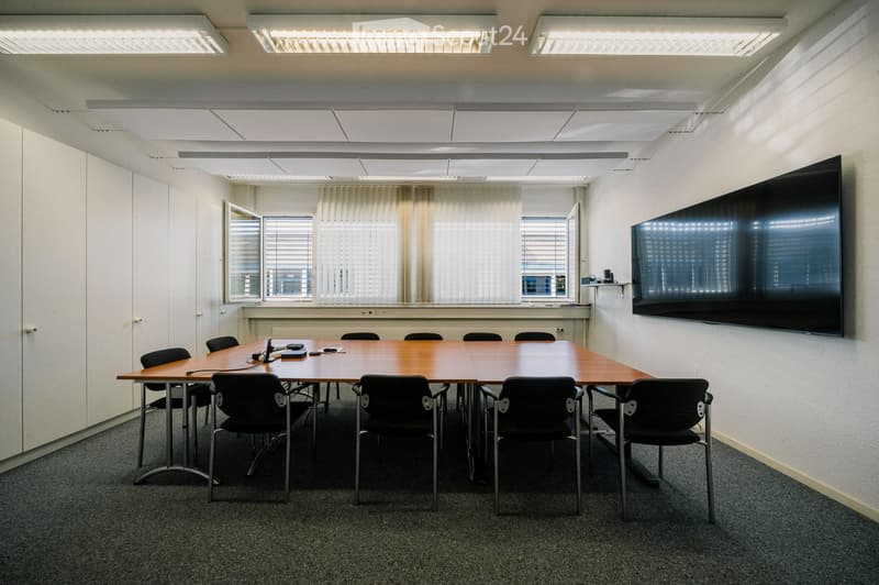 Conference room with a large wooden table, black chairs, and a large flat-screen TV mounted on the wall. The room has white walls, carpeted flooring, and large windows with blinds.