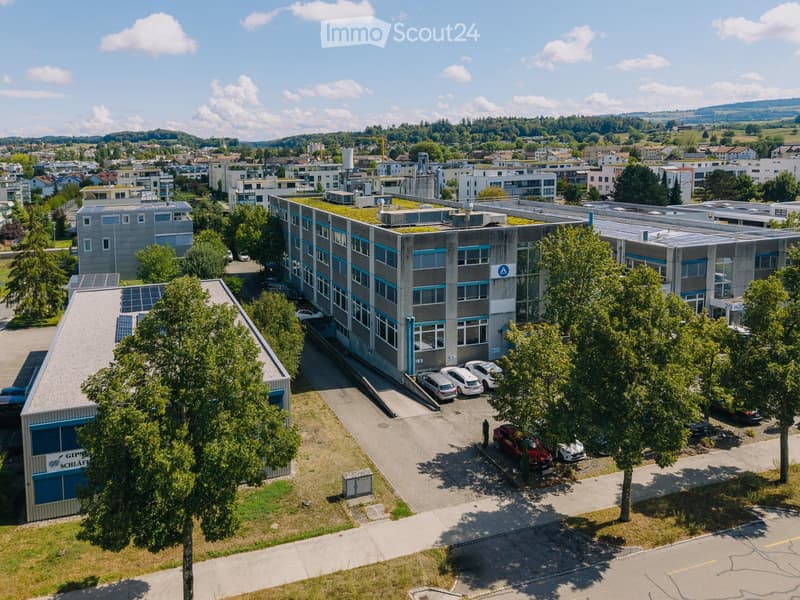 Aerial view of a modern industrial building with a green roof, surrounded by trees and parked cars in a suburban area.