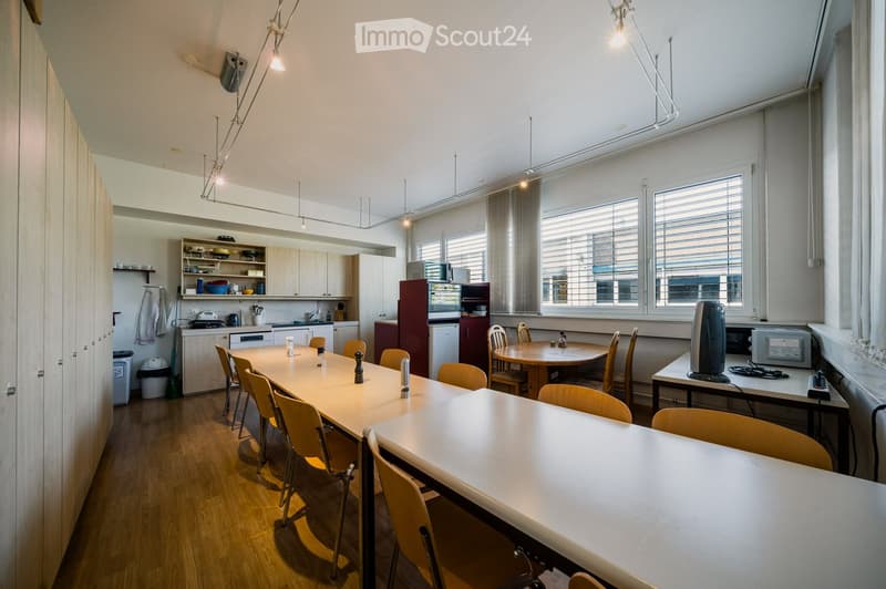 Bright, open-plan room with a long dining table and chairs, a smaller round table with chairs, a kitchen area with cabinets, a refrigerator, microwave, and a counter, and a workspace with a screen and other items.