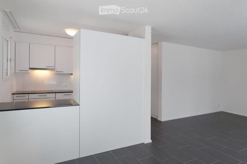 Empty room with white walls, dark tiled floor, and a white kitchen area with cabinets and a countertop visible in the background.