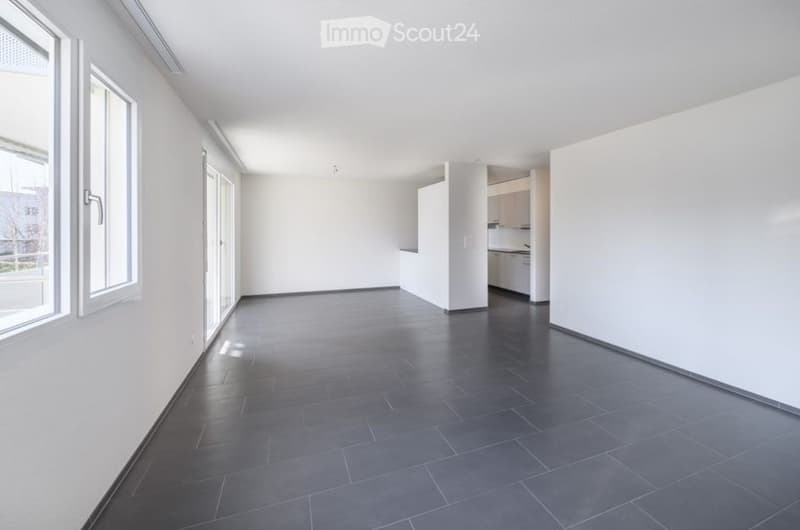 Empty room with white walls, dark tiled floor, large windows, and a small kitchen area with cabinets and appliances visible in the background.