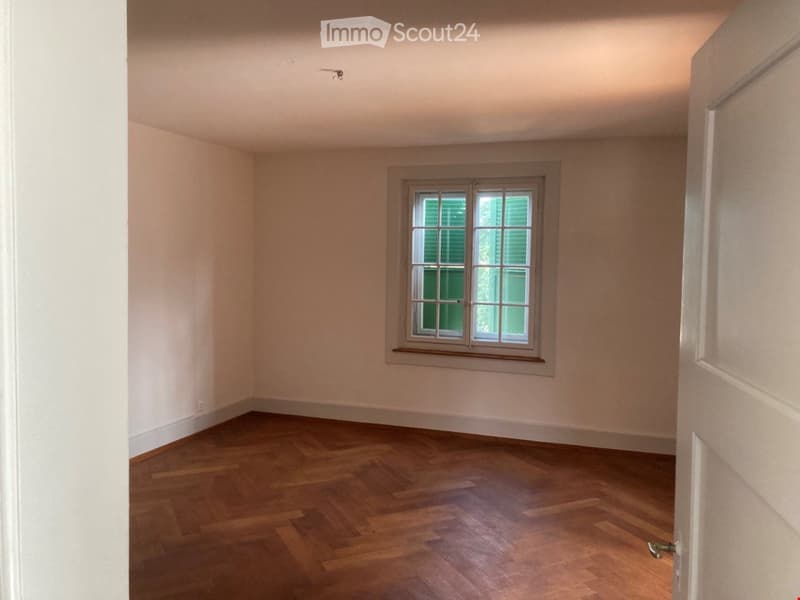 Empty room with wooden herringbone flooring, white walls, and a single window with green shutters.