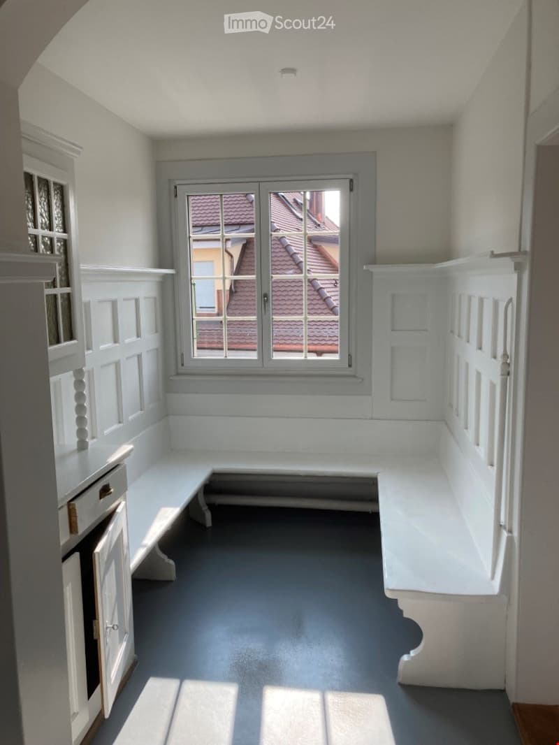 Empty room with white paneled walls, built-in bench seating, dark grey floor, and a large window showing a roofed building outside.