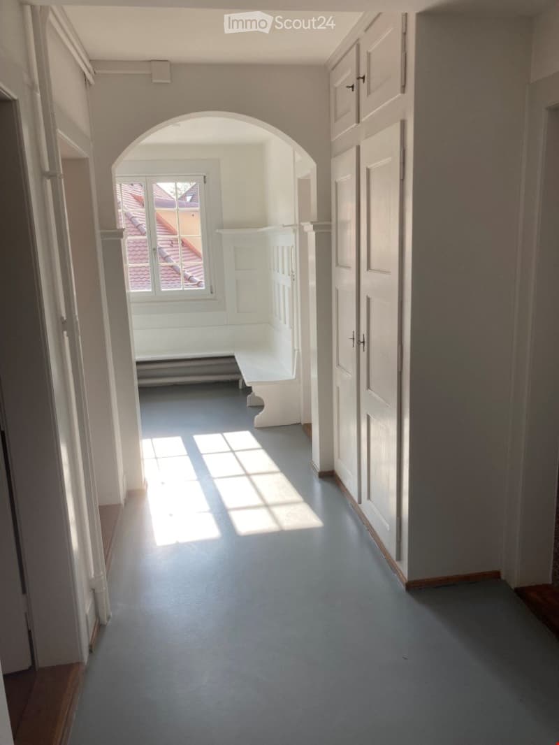 Empty hallway with white walls, grey carpeted floor, and an arched opening leading to a small seating area with a bench and window.