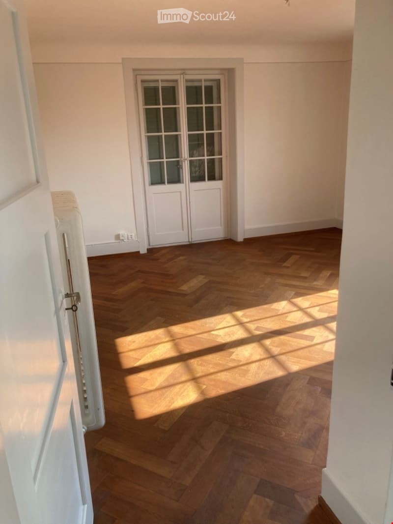 Empty room with wooden herringbone flooring, white walls, and a white double door with glass panels.
