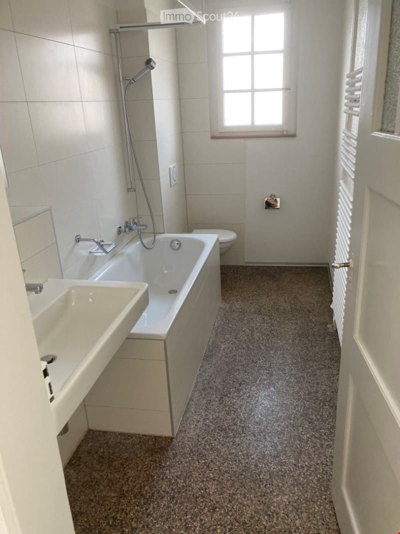 Modern bathroom with a bathtub, sink, toilet, and showerhead. The room features white tiled walls and a grey speckled floor.